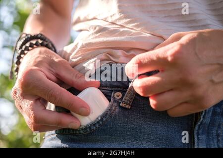 Woman hand puts a case with wireless headphones in the pocket of denim shorts, outdoors. Close-up. Stock Photo