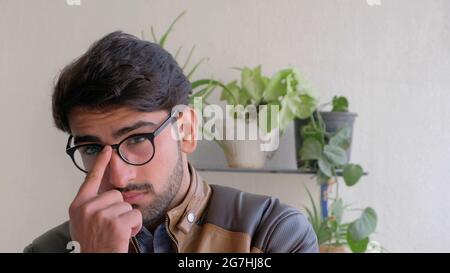 Young Indian male in eyeglasses wearing a brown leather jacket with houseplants in the background Stock Photo