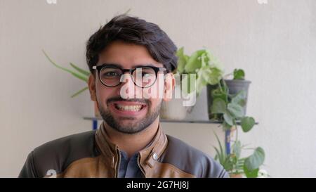 Happy young Indian male in eyeglasses wearing a leather jacket with houseplants in the background Stock Photo