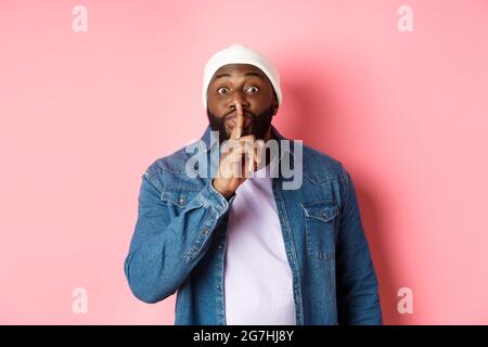 Handsome young black guy in beanie and denim shirt prepare surprise, hushing softly and looking at camera, asking keep quiet, pink background Stock Photo