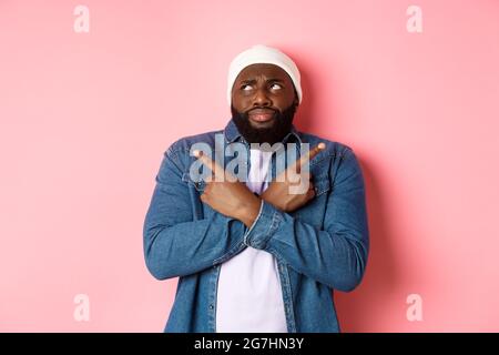 Confused Black man with beard, making choice, pointing fingers sideways and looking puzzled, standing over pink background Stock Photo
