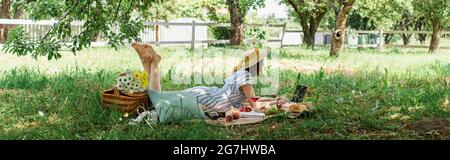 Woman in summer dress using laptop near food, wine and flowers in park, banner Stock Photo