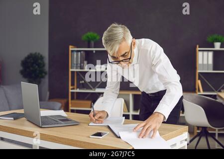 Senior businessman standing by the desk in his office and taking notes in his notebook Stock Photo