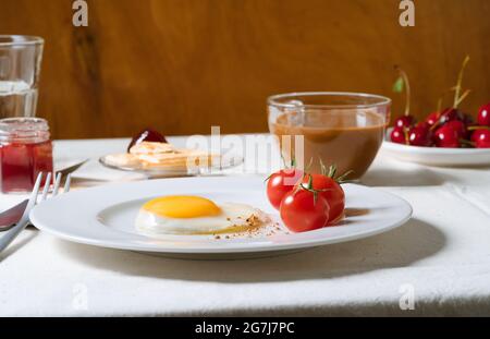 Dining table set for breakfast. Coffee, milk, fried egg, cherry tomatoes, fruit jam, sweet cherries and water on linen tablecloth Stock Photo
