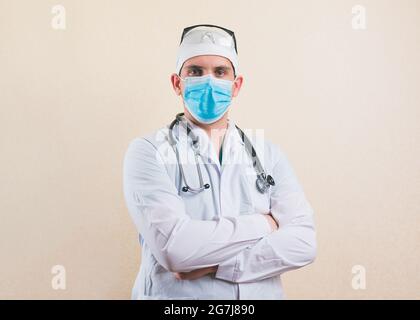 Young doctor in white suit, protective glasses, stethoscope and protective breathing mask with his hands on his chest isolated. Stock Photo