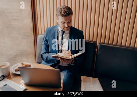 Young bearded businessman sitting in cafe and writing down notes during online webinar Stock Photo