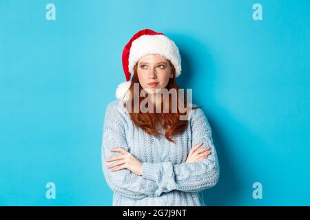 Winter holidays and Christmas Eve concept. Thoughtful redhead woman in Santa hat and sweater, looking left and pondering, making xmas plans, standing Stock Photo