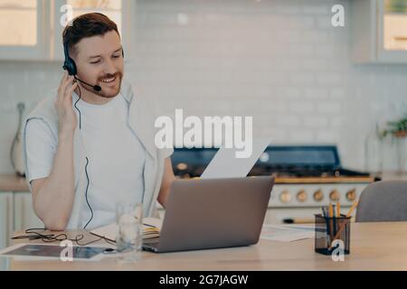 Indoor shot of busy European man has video courses watches webinar listens lecture studies online wears headphones focused at laptop screen dressed Stock Photo