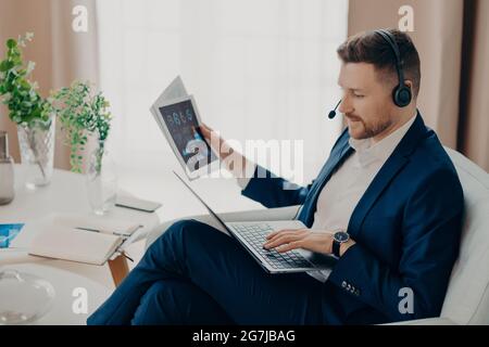 Male financial expert looks through papers with graphics dressed in formal clothes checks information on laptop computer wears headset poses against Stock Photo