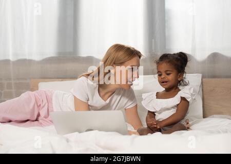 surprised mother and adopted african american girl watching movie on laptop in bedroom Stock Photo