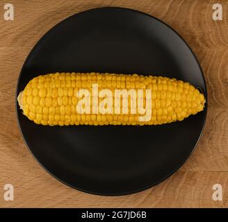 Ripe corn in a head of cabbage peeled from the husk lies on a black plate, on a wooden board Stock Photo