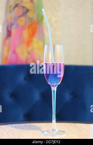 a champagne glass standing on a table with a straw and a drink of a purple hue on the background of a blue chair Stock Photo