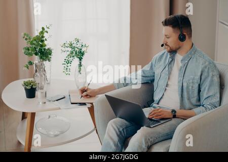 Attentive guy making notes in notebook during an online lesson at home Stock Photo