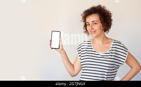 A curly haired Caucasian woman poses next to her mobile phone, looking at camera Stock Photo