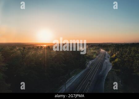 Shiny rails at turn of railway among coniferous forest at sunset on a clear summer day. Picturesque landscape: rails of railway against background of the Sun setting over the horizon in summer. Stock Photo