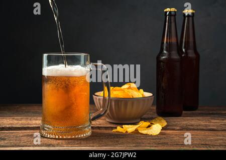 Pouring beer into a glass against the background of chips and beer bottles. Stock Photo