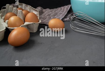 Fresh eggs in cardboard egg tray with whisk, plate and towel on dark background Stock Photo