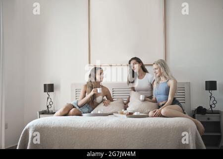 Female friends having coffee and talking while sitting on a bed in a bedroom Stock Photo
