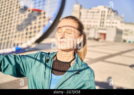 Portrait of attractive middle aged sportswoman holding tennis racket, standing with eyes closed, ready for morning workout outdoors Stock Photo