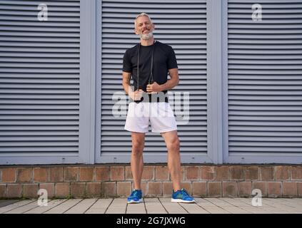 Middle aged athletic man in sportswear holding skipping rope while standing outdoors over grey background Stock Photo