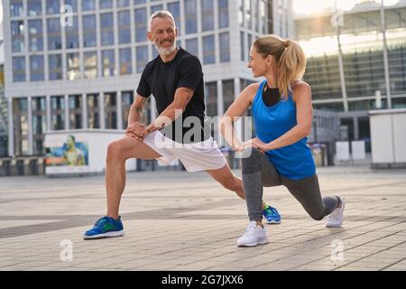 Active sporty middle aged couple, man and woman stretching legs, preparing for running together in the city on a summer day Stock Photo