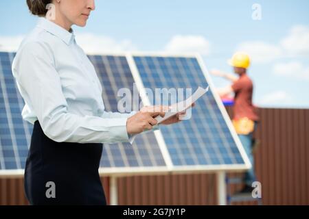 Serious businesswoman reading document when mounter installing solar panel in background Stock Photo