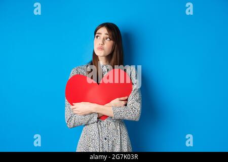 Valentines day. Sad heartbroken girl sobbing, looking aside at empty space and crying upset, hugging big red heart, feeling lonely without lover Stock Photo