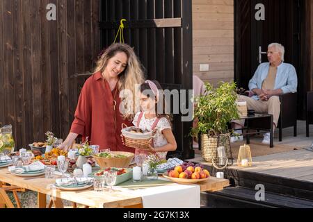 Pretty little girl putting basket of bread on dinner table helping her mom to serve table for family event Stock Photo