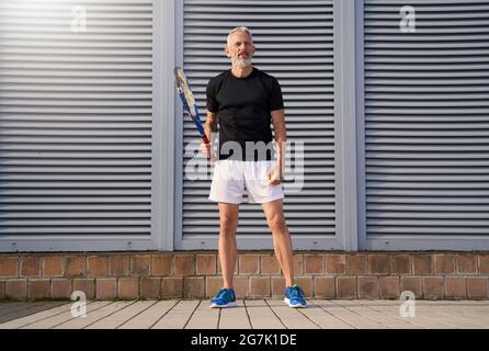 Fit mature man in sportswear holding tennis racket and looking concentrated while standing outdoors over gray background Stock Photo