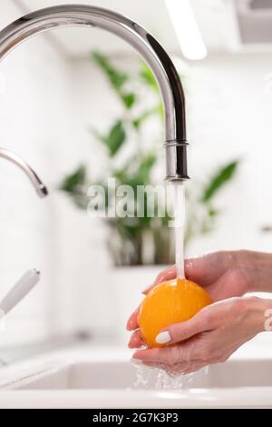 Female washing orange fruit in fresh water from kitchen sink crane. Hygiene, healthcare and safety Stock Photo