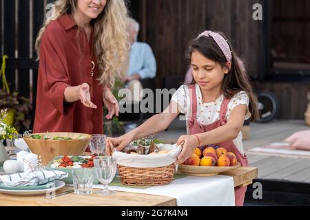 Girl helping mother with serving dinner table and putting basket of bread on table Stock Photo