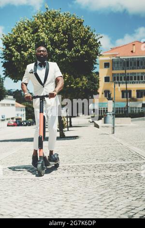 A vertical shot of a dapper adult bearded bald black senior in eyeglasses and a white fancy summer costume is holding in hands the handlebars of the e Stock Photo