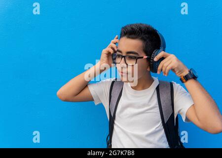 Caucasian boy with backpack, putting on headphones leaning against a blue wall Stock Photo