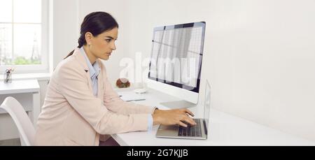 Young businesswoman sitting at office desk and working on desktop and laptop computers Stock Photo