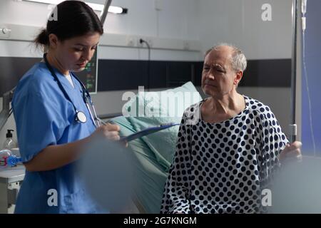Senior sick man in hospital room discussing with nurse about treatment during consultation. Medical staff with stethoscope writing notes on clipboard preparing patient for surgery. Stock Photo