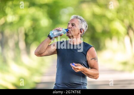 Handsome elderly man rehydrates after a run outdoors in the nature by drinking water from a bottle. Stock Photo