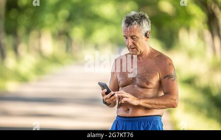 Elderly but fit man wears earphones and checks his mobile phone post-workout outdoors. Stock Photo