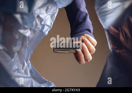 Hand tosses comb into a trash can. Man holding old black plastic comb over garbage basket. View from below. Indoors. Stock Photo
