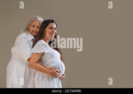 A YOUNG PREGNANT WOMAN WITH MOTHER SMILING AND POSING WHILE EMBRACING BABY BUMP Stock Photo
