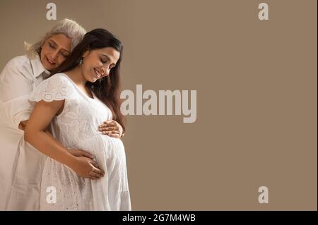 A SENIOR WOMAN AND PREGNANT DAUGHTER HAPPILY WRAPPING HANDS AROUND BABY BUMP Stock Photo