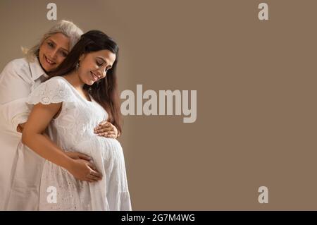 A SENIOR WOMAN WITH PREGNANT DAUGHTER BESIDES LOOKING AT CAMERA AND SMILING AND EMBRACING BABY BUMP Stock Photo