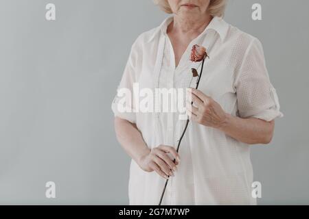 Upset elderly woman in white dressed hold withered single one dry old rose flower Stock Photo