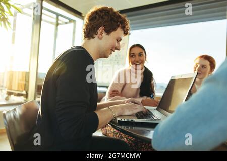 Male student sitting at table and doing project on laptop with classmates sitting by in college. Boy using laptop while doing group studies with frien Stock Photo