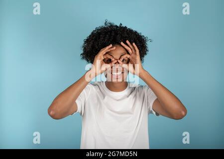 Smiling African American young man in his 20s in basic casual white t-shirt with hands near eyes, imitating glasses or binoculars isolated on pastel b Stock Photo