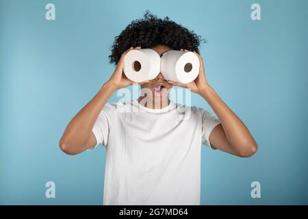 Afro-haired young man in basic white t-shirt looks astonishingly through toilet paper rolls, simulating binoculars. Isolated on blue background. Stock Photo