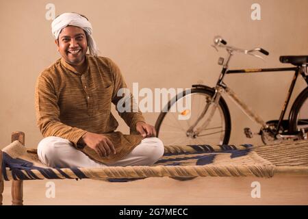 A TURBANED VILLAGER HAPPILY SITTING ON A COT AND LOOKING AT CAMERA Stock Photo