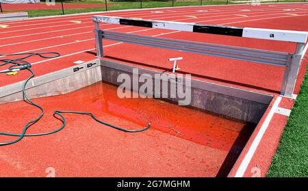 Empty steeplechase pit being filled up with water on a track from a green hose for for runners to compete. Stock Photo