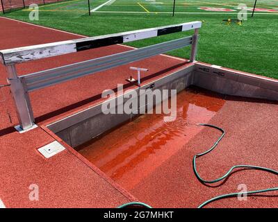 A red track and field steeplechase pit is being filled with water by a green hose so track runners can jump into it during their running race. Stock Photo