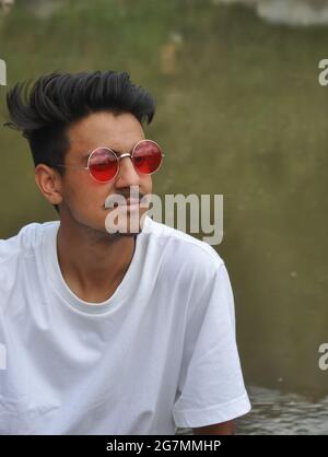 Closeup of a thoughtful young guy sitting beside of river with looking sideways, A Indian guy with wearing white t-shirt and red sunglasses Stock Photo