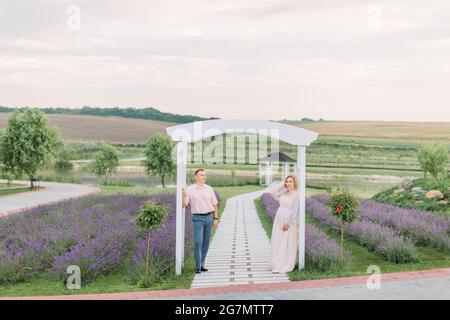 Happy together. Beautiful stylish middle aged couple in the lavender field, standing and looking away, leaning on the big white wooden arch Stock Photo